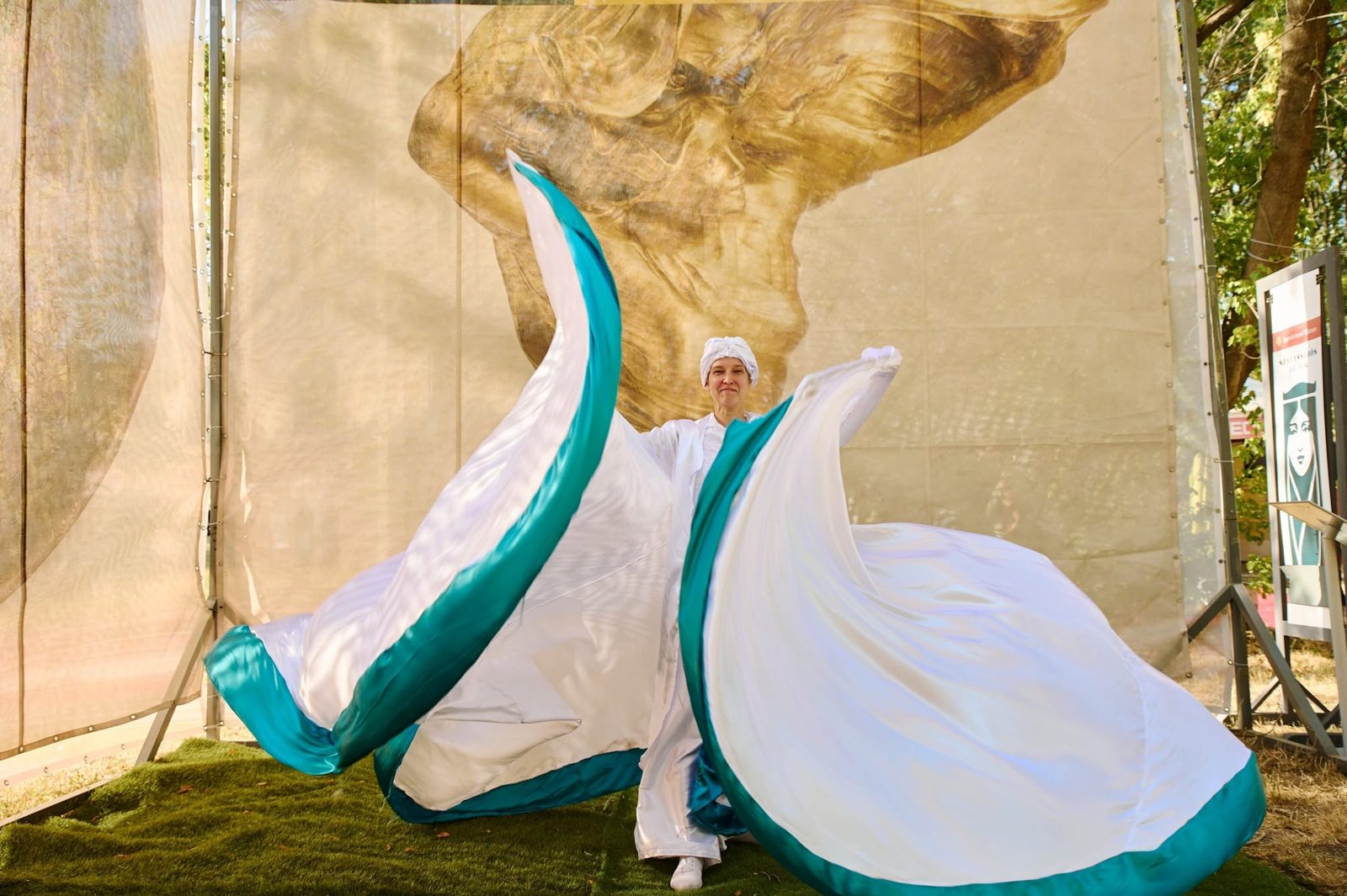 Loie Fuller a Sziget Fesztiválon: táncos fehér-türkiz selyemszárnyakkal az Iparművészeti Múzeum Loie Fuller-szobrot ábrázoló hátterénél